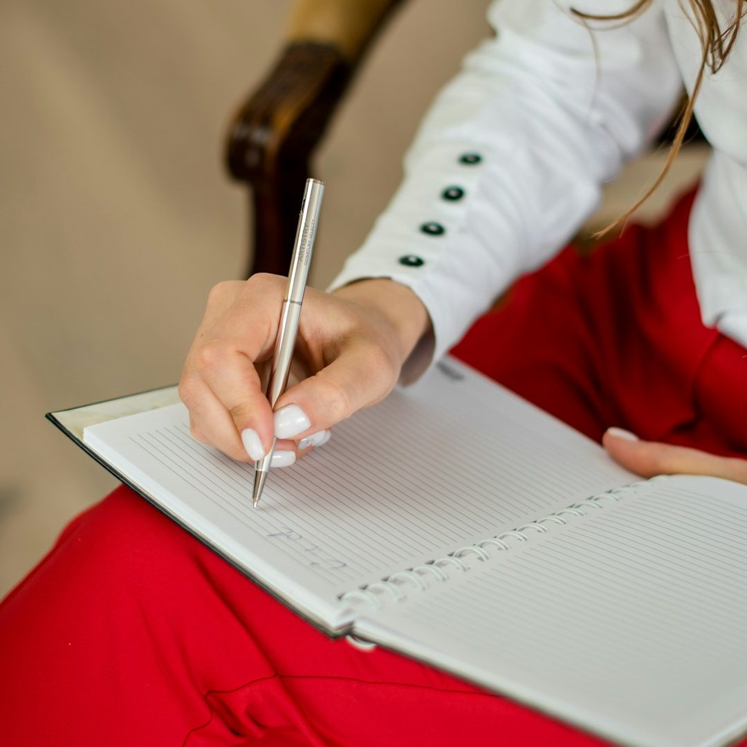 Dossier médiation Une main écrivant dans un carnet avec un stylo, portée par une personne en jupe rouge.